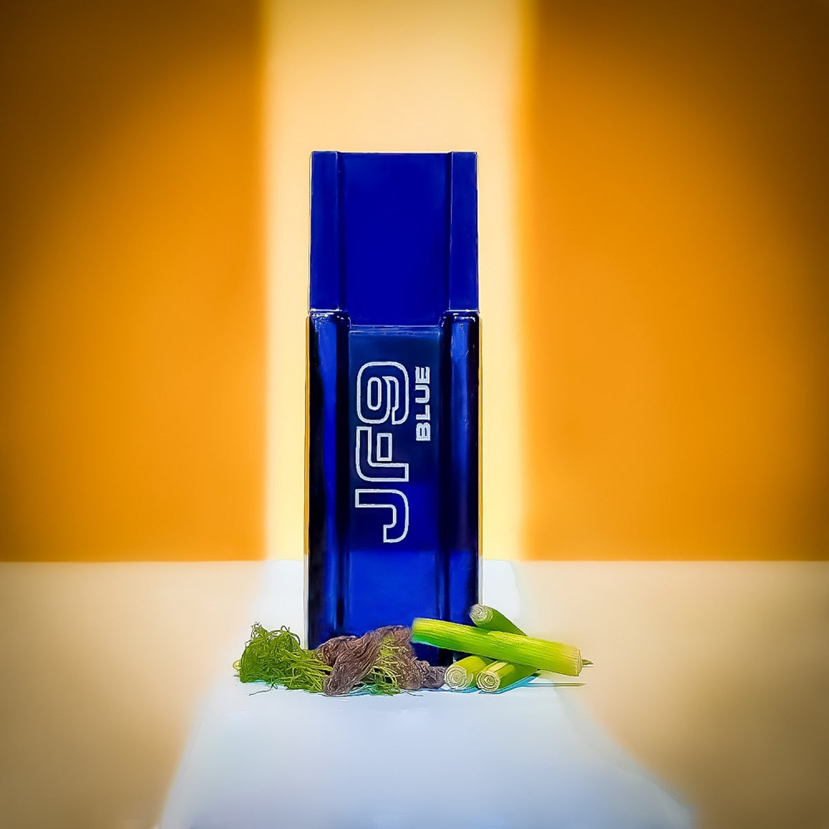 Blue JF9 perfume bottle with green leaves on a white surface against an orange background